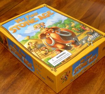 Board game box for "My First Stone Age," featuring colorful illustrations of early humans and a woolly mammoth, designed for ages 2-4, suitable for 2-4 players, with a playtime of 15 minutes.