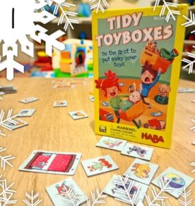 Colorful board game "Tidy Toyboxes" by HABA, featuring playful illustrations on the box and scattered game cards on a wooden table, promoting fun and organization for children.