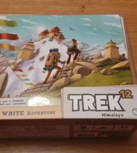 Box cover of the board game "Trek 12: Himalaya," featuring vibrant artwork of two adventurers trekking through a mountainous landscape with prayer flags and a distant temple. The design highlights the game's theme of exploration and adventure in the Himalayas.