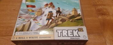 Box cover of the board game "Trek 12: Himalaya," featuring vibrant artwork of two adventurers trekking through a mountainous landscape with prayer flags and a distant temple. The design highlights the game's theme of exploration and adventure in the Himalayas.