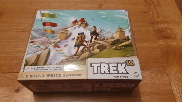 Box cover of the board game "Trek 12: Himalaya," featuring vibrant artwork of two adventurers trekking through a mountainous landscape with prayer flags and a distant temple. The design highlights the game's theme of exploration and adventure in the Himalayas.