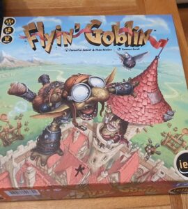 Image of the board game "Flyin' Goblin" featuring colorful artwork of a goblin in goggles flying above a medieval village, with game components visible on the box. The game is designed for 2-4 players and offers a playful theme of goblin adventures.