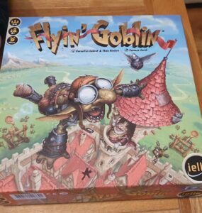 Image of the board game "Flyin' Goblin" featuring colorful artwork of a goblin in goggles flying above a medieval village, with game components visible on the box. The game is designed for 2-4 players and offers a playful theme of goblin adventures.