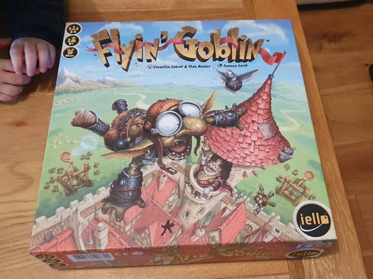 Image of the board game "Flyin' Goblin" featuring colorful artwork of a goblin in goggles flying above a medieval village, with game components visible on the box. The game is designed for 2-4 players and offers a playful theme of goblin adventures.