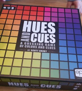 Hues and Cues board game box featuring a colorful grid design, designed by Scott Brady, displayed on a wooden table with hands partially visible, emphasizing the interactive nature of the game focused on color guessing and clues.