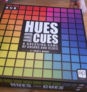 Hues and Cues board game box featuring a colorful grid design, designed by Scott Brady, displayed on a wooden table with hands partially visible, emphasizing the interactive nature of the game focused on color guessing and clues.