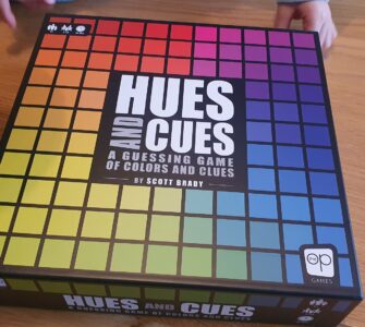 Hues and Cues board game box featuring a colorful grid design, designed by Scott Brady, displayed on a wooden table with hands partially visible, emphasizing the interactive nature of the game focused on color guessing and clues.