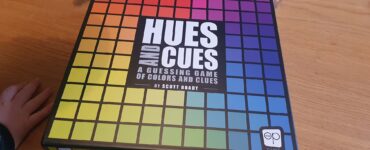 Hues and Cues board game box featuring a colorful grid design, designed by Scott Brady, displayed on a wooden table with hands partially visible, emphasizing the interactive nature of the game focused on color guessing and clues.