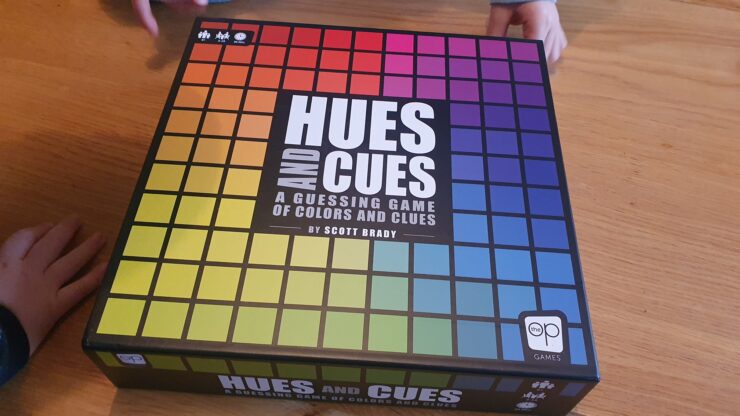 Hues and Cues board game box featuring a colorful grid design, designed by Scott Brady, displayed on a wooden table with hands partially visible, emphasizing the interactive nature of the game focused on color guessing and clues.