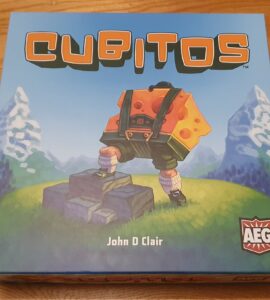 Box cover of the board game "Cubitos" by John D. Clair, featuring a character with a cheese-themed backpack standing on rocks, set against a vibrant landscape with mountains and greenery. The game's logo and publisher's emblem are prominently displayed.