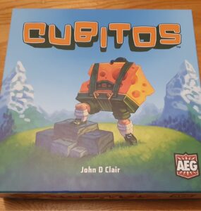 Box cover of the board game "Cubitos" by John D. Clair, featuring a character with a cheese-themed backpack standing on rocks, set against a vibrant landscape with mountains and greenery. The game's logo and publisher's emblem are prominently displayed.