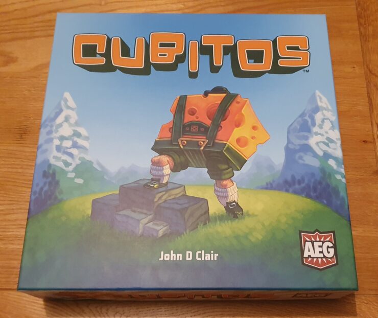 Box cover of the board game "Cubitos" by John D. Clair, featuring a character with a cheese-themed backpack standing on rocks, set against a vibrant landscape with mountains and greenery. The game's logo and publisher's emblem are prominently displayed.