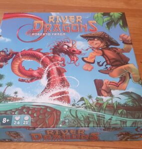Colorful board game box for "River Dragons" by Roberto Fraga, featuring a boy in a straw hat and a dragon amidst a river scene with tropical plants. Game details indicate it's suitable for ages 8+, for 2-6 players, with a playtime of approximately 25 minutes.