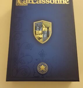 Box cover of the board game Carcassonne by Klaus-Jürgen Wrede, featuring a gold and blue shield design, intricate patterns, and a meeple symbol, set against a dark background.