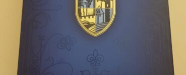 Box cover of the board game Carcassonne by Klaus-Jürgen Wrede, featuring a gold and blue shield design, intricate patterns, and a meeple symbol, set against a dark background.