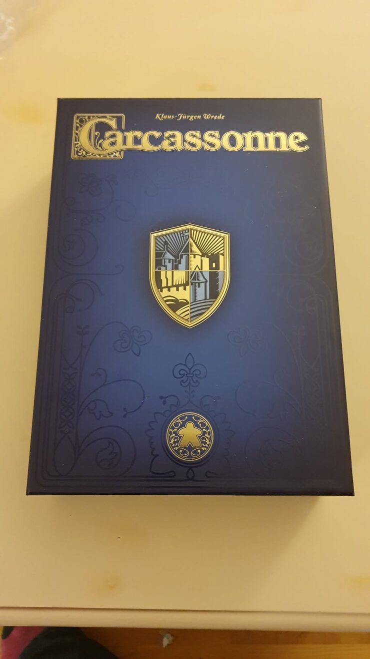 Box cover of the board game Carcassonne by Klaus-Jürgen Wrede, featuring a gold and blue shield design, intricate patterns, and a meeple symbol, set against a dark background.