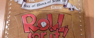 Deluxe Edition of the board game "Roll for It!" by Calliope Games, featuring an ornate tin design with an illustration of a woman and the tagline "Roll It! Match It! Score It!" on the lid, placed on a wooden surface.