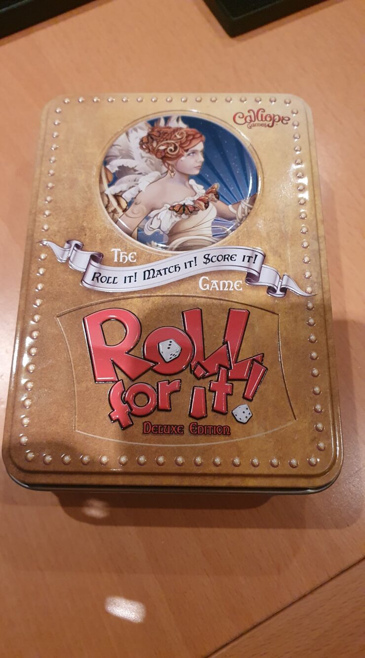 Deluxe Edition of the board game "Roll for It!" by Calliope Games, featuring an ornate tin design with an illustration of a woman and the tagline "Roll It! Match It! Score It!" on the lid, placed on a wooden surface.