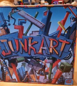 Colorful board game box of "Junk Art," featuring dynamic artwork of characters building structures with various junk pieces, set on a wooden table.