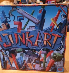 Colorful board game box of "Junk Art," featuring dynamic artwork of characters building structures with various junk pieces, set on a wooden table.