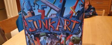 Colorful board game box of "Junk Art," featuring dynamic artwork of characters building structures with various junk pieces, set on a wooden table.