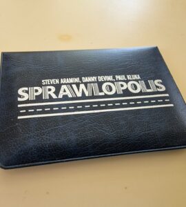 Black cover of the board game "Sprawlopolis" by Steven Aramini, Danny Devine, and Paul Kluka, placed on a beige surface.