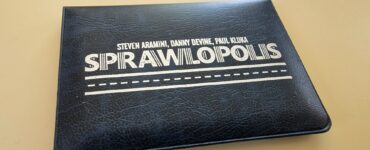 Black cover of the board game "Sprawlopolis" by Steven Aramini, Danny Devine, and Paul Kluka, placed on a beige surface.