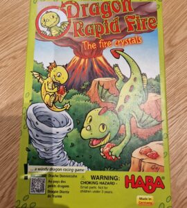 Illustration of the board game "Dragon Rapid Fire" by HABA, featuring colorful cartoon dragons and a volcano. The game is designed for children, emphasizing fun and adventure in a windy dragon racing theme. Contains a warning about small parts being a choking hazard for children under three years. Made in Germany.