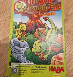 Illustration of the board game "Dragon Rapid Fire" by HABA, featuring colorful cartoon dragons and a volcano. The game is designed for children, emphasizing fun and adventure in a windy dragon racing theme. Contains a warning about small parts being a choking hazard for children under three years. Made in Germany.