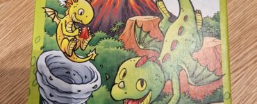 Illustration of the board game "Dragon Rapid Fire" by HABA, featuring colorful cartoon dragons and a volcano. The game is designed for children, emphasizing fun and adventure in a windy dragon racing theme. Contains a warning about small parts being a choking hazard for children under three years. Made in Germany.