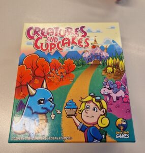 Colorful board game box for "Creatures and Cupcakes" featuring a playful dragon and a girl holding a cupcake, set against a whimsical landscape with a cupcake castle and cute creatures. Designed by Daryl Andrews and Adrian Adamescu, published by Social Sloth Games.