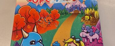 Colorful board game box for "Creatures and Cupcakes" featuring a playful dragon and a girl holding a cupcake, set against a whimsical landscape with a cupcake castle and cute creatures. Designed by Daryl Andrews and Adrian Adamescu, published by Social Sloth Games.