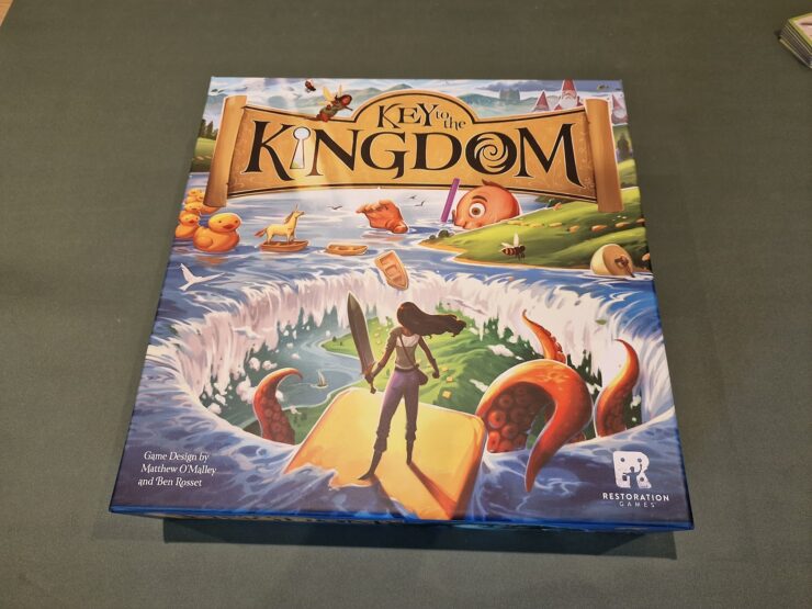 Board game box of "Key to the Kingdom," featuring colorful artwork with a character standing on a yellow path, surrounded by whimsical elements like rubber ducks and an octopus, designed by Matthew O'Malley and Ben Rosset, published by Restoration Games.
