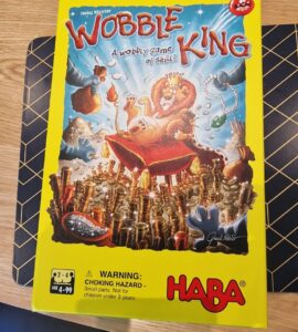 Colorful board game box for "Wobble King" by HABA, featuring playful illustrations of animals and a lion wearing a crown. The box highlights the game's theme of skill and balance, with a warning about small parts for children under 3 years. Suitable for 2-4 players aged 4 to 99.