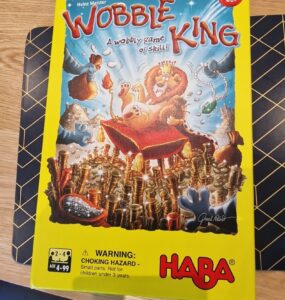 Colorful board game box for "Wobble King" by HABA, featuring playful illustrations of animals and a lion wearing a crown. The box highlights the game's theme of skill and balance, with a warning about small parts for children under 3 years. Suitable for 2-4 players aged 4 to 99.