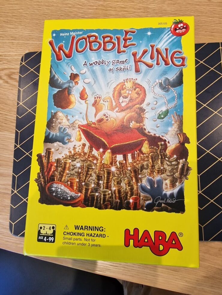 Colorful board game box for "Wobble King" by HABA, featuring playful illustrations of animals and a lion wearing a crown. The box highlights the game's theme of skill and balance, with a warning about small parts for children under 3 years. Suitable for 2-4 players aged 4 to 99.