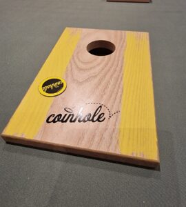 Wooden coinhole game board featuring a circular hole and vibrant yellow accents, placed on a green surface, with a sticker logo visible.