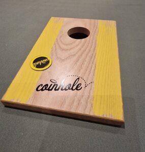 Wooden coinhole game board featuring a circular hole and vibrant yellow accents, placed on a green surface, with a sticker logo visible.