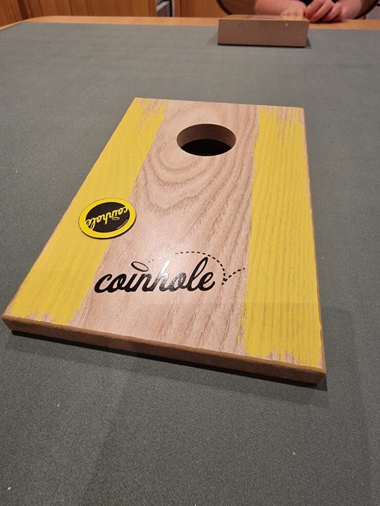 Wooden coinhole game board featuring a circular hole and vibrant yellow accents, placed on a green surface, with a sticker logo visible.