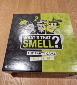 Colorful game box for "What's That Smell?" featuring humorous illustrations and the tagline "The Party Game That Stinks!" Suitable for ages 14 and up, for 2 to 6 players.