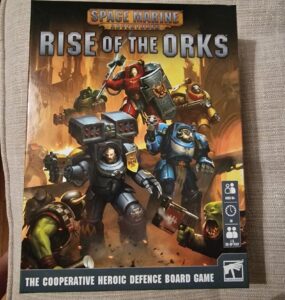 Board game box cover for "Space Marine Adventures: Rise of the Orks," featuring heroic Space Marines battling Orks. The game is designed for 1-5 players, ages 8 and up, and focuses on cooperative defense gameplay.