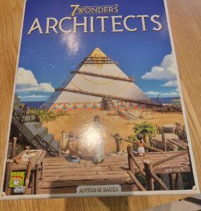 Box cover of the board game "7 Wonders: Architects" by Antoine Bauza, featuring a colorful illustration of a pyramid under construction with characters engaged in architectural activities, set against a beach backdrop. The game is produced by Repos Production.