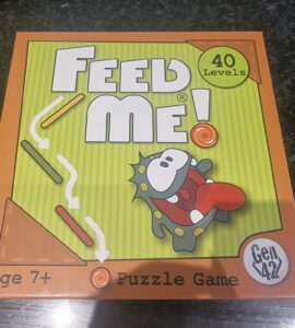 Colorful box of the "Feed Me!" puzzle game featuring a cartoon character, labeled for ages 7 and up, with 40 levels of gameplay. Ideal for family fun and cognitive development.