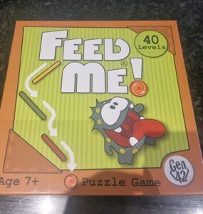 Colorful box of the "Feed Me!" puzzle game featuring a cartoon character, labeled for ages 7 and up, with 40 levels of gameplay. Ideal for family fun and cognitive development.