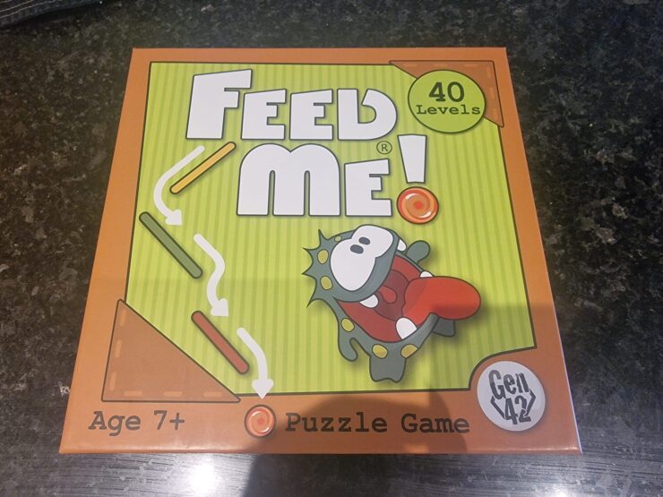 Colorful box of the "Feed Me!" puzzle game featuring a cartoon character, labeled for ages 7 and up, with 40 levels of gameplay. Ideal for family fun and cognitive development.