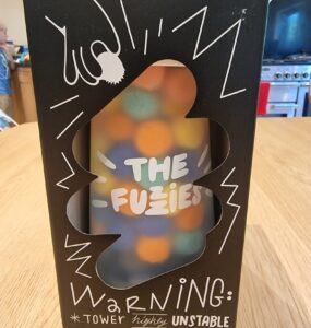 Boxed game "The Fuzies" displayed on a wooden table, featuring a colorful tower design and a warning label indicating it is "highly unstable." The packaging includes playful graphics and text, emphasizing the game's fun and chaotic nature.