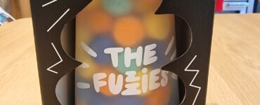 Boxed game "The Fuzies" displayed on a wooden table, featuring a colorful tower design and a warning label indicating it is "highly unstable." The packaging includes playful graphics and text, emphasizing the game's fun and chaotic nature.