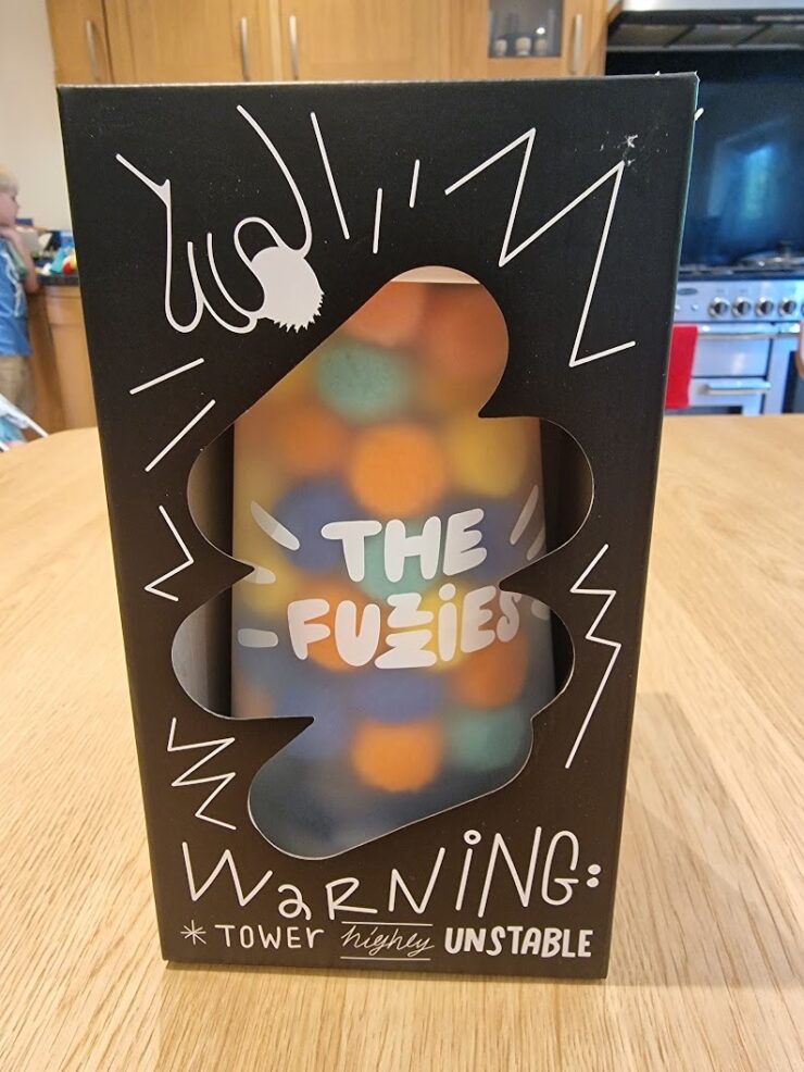 Boxed game "The Fuzies" displayed on a wooden table, featuring a colorful tower design and a warning label indicating it is "highly unstable." The packaging includes playful graphics and text, emphasizing the game's fun and chaotic nature.