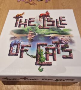 Board game box for "The Isle of Cats" featuring colorful illustrations of cats and a ship surrounded by a lush landscape, placed on a wooden table.