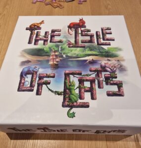 Board game box for "The Isle of Cats" featuring colorful illustrations of cats and a ship surrounded by a lush landscape, placed on a wooden table.